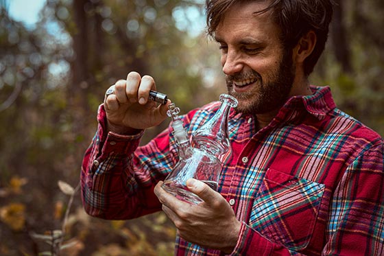 Man in red checkered shirt lighting up a glass bong in the outdoors.