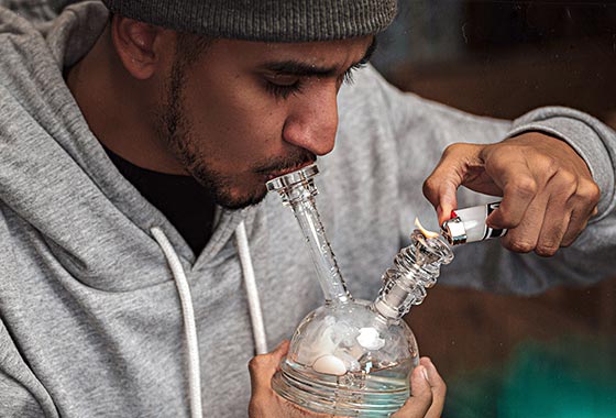 Man wearing hoodie and hat, lighting up a glass marijuana pipe.